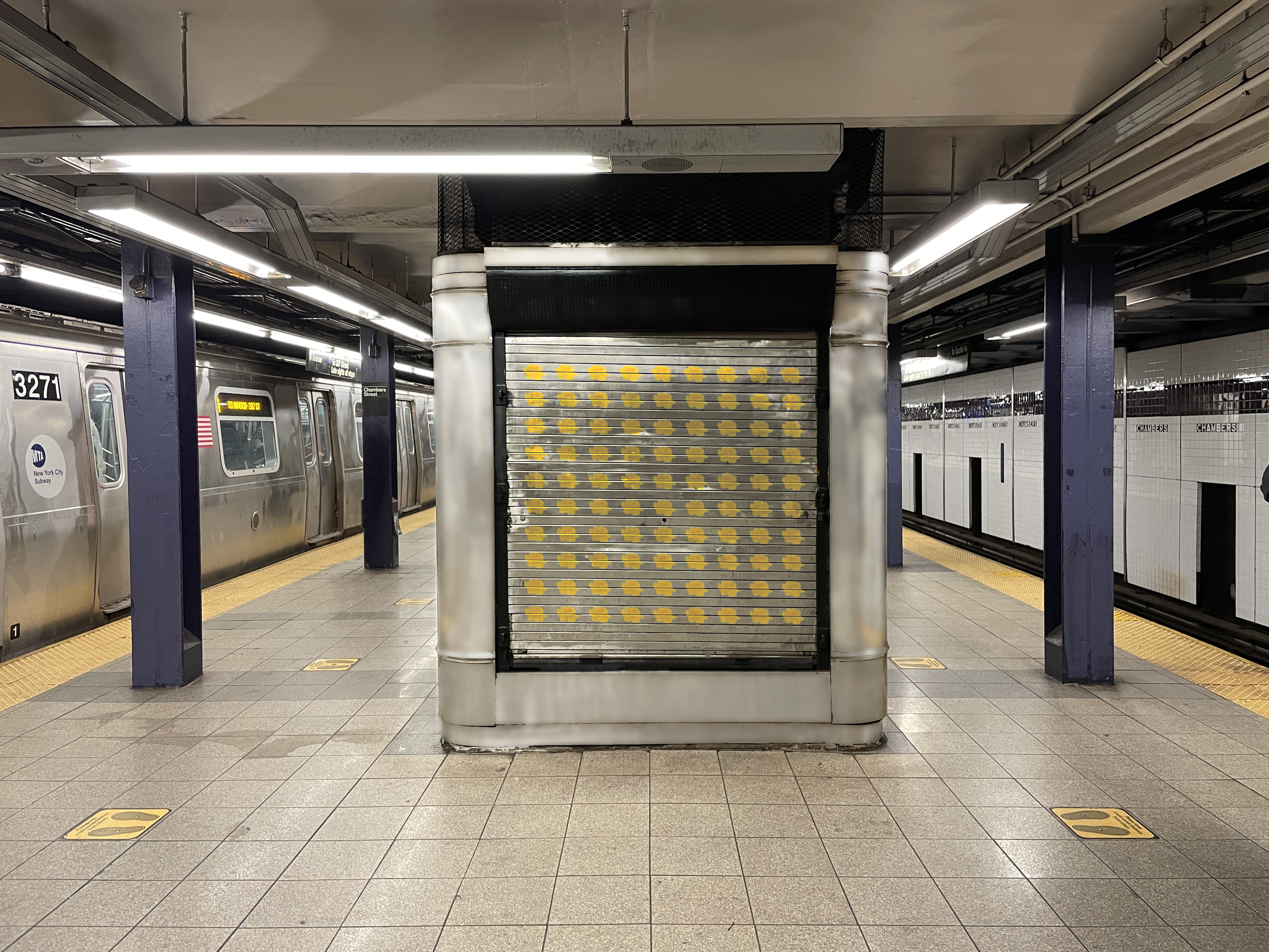 A photo of a closed subway platform storefront unit with a pattern of yellow flowers painted on the stainless steel roll-up door. The subway station signs read “Chambers Street.”